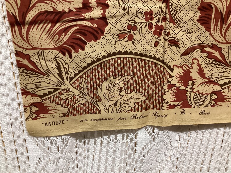 May include: Close-up of a beige fabric with a brown and red floral pattern. The fabric has a label that reads "ANDUZE" en imprim&eacute; par Roland Sigrist Paris.