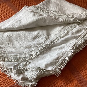 May include: A folded white blanket with a textured surface and fringe trim. The blanket features a diamond and floral pattern. The blanket is resting on an orange waffle-weave surface.