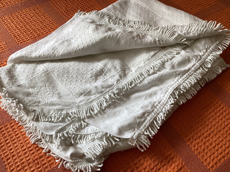 May include: A folded white blanket with a textured surface and fringe trim. The blanket features a diamond and floral pattern. The blanket is resting on an orange waffle-weave surface.