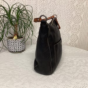 Pu&ograve; includere: Borsa a tracolla in pelle nera con finitura strutturata e cuciture bianche. La borsa ha una singola tasca esterna e un manico in pelle marrone. Una piccola pianta in vaso &egrave; sullo sfondo.