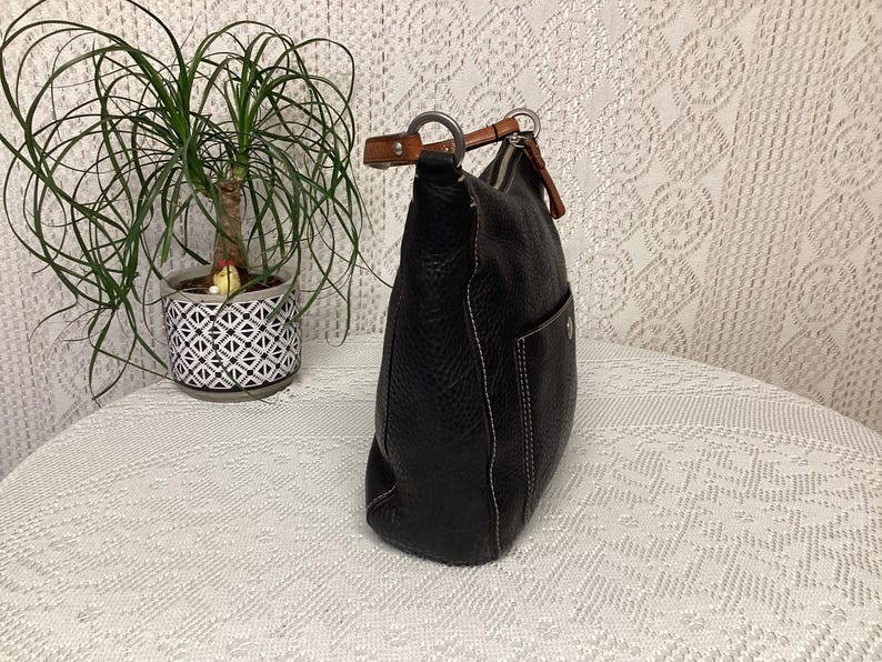 Pu&ograve; includere: Borsa a tracolla in pelle nera con finitura strutturata e cuciture bianche. La borsa ha una singola tasca esterna e un manico in pelle marrone. Una piccola pianta in vaso &egrave; sullo sfondo.
