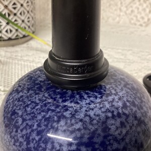 May include: A close-up of a blue and white speckled glass object with a black cylindrical top. The base of the top has the word "Kneeb&euml;rger" printed on it. A black decorative object is in the background.