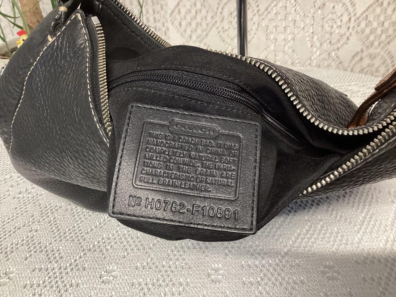 Pu&ograve; includere: Borsa Coach in pelle nera con chiusura a zip e cuciture bianche. La borsa ha una superficie strutturata e un'etichetta in pelle nera all'interno con testo e il numero H0782-F10891.
