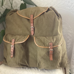 May include: A vintage green canvas rucksack with brown leather straps and buckles. The rucksack has two large front pockets with additional buckles.
