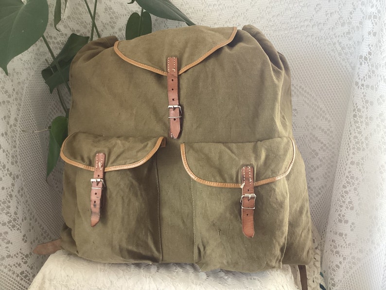May include: A vintage green canvas backpack with brown leather straps and buckles. The backpack has two large front pockets with additional buckles.