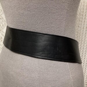 May include: A wide, black leather belt with a curved design. The belt is displayed on a mannequin, showcasing its shape and texture. The belt appears to be made of a sturdy material.