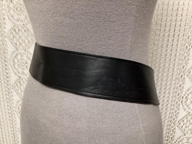 May include: A wide, black leather belt with a curved design. The belt is displayed on a mannequin, showcasing its shape and texture. The belt appears to be made of a sturdy material.