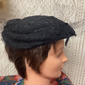 May include: A black lace hat with a veil. The hat is perched on a mannequin head.