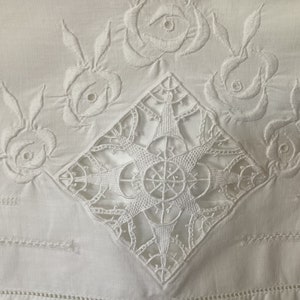 May include: White cotton fabric with intricate embroidered designs. The center features a large, geometric, star-shaped pattern. The corners are decorated with smaller floral motifs.