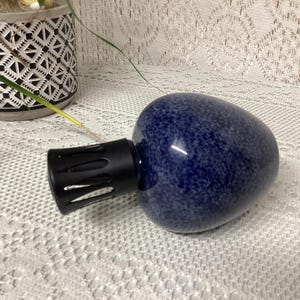 May include: A dark blue essential oil diffuser with a speckled pattern and a black nozzle. The diffuser is egg-shaped and sits on a white lace tablecloth. A decorative pot with a geometric pattern is in the background.