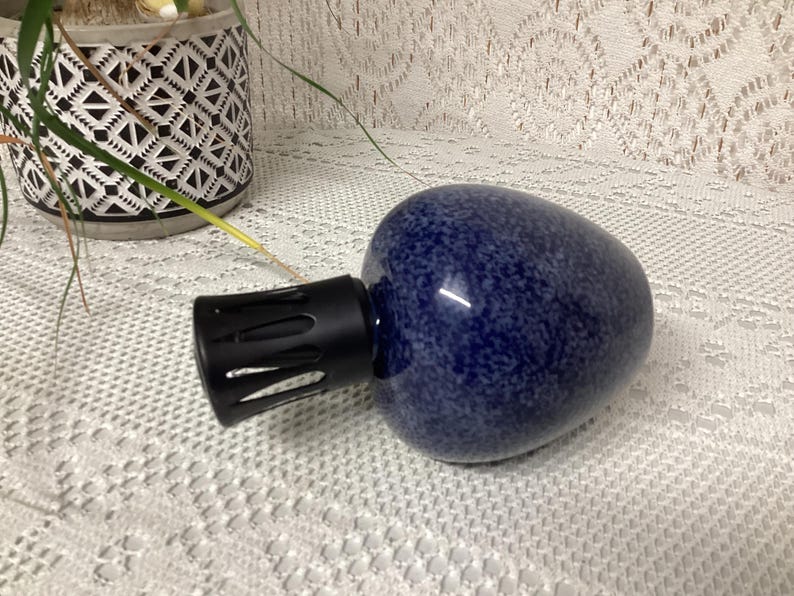 May include: A dark blue essential oil diffuser with a speckled pattern and a black nozzle. The diffuser is egg-shaped and sits on a white lace tablecloth. A decorative pot with a geometric pattern is in the background.