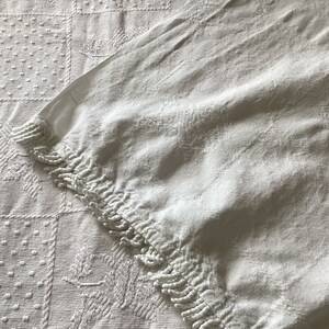 May include: Close-up of a white textile with a textured surface. The fabric features a detailed pattern of squares, floral designs, and dotted textures. The edge of the textile has a fringe detail. The overall aesthetic is vintage and elegant.