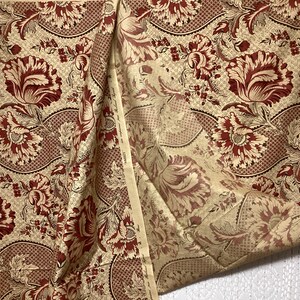 May include: A beige fabric with a repeating pattern of red and gold floral designs. The fabric has a subtle texture and a slightly distressed look.