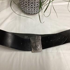 May include: A wide black leather belt with a silver-colored buckle featuring a carved face design. The belt has multiple holes for adjustment and is displayed on a white surface.