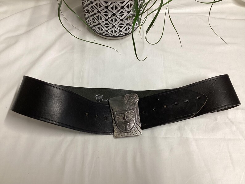 May include: A wide black leather belt with a silver-colored buckle featuring a carved face design. The belt has multiple holes for adjustment and is displayed on a white surface.