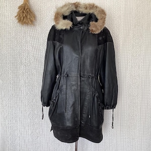 Op de afbeelding: Zwarte leren jas met een met bont afgezette capuchon. De jas heeft een taille met trekkoord, zakken en suède schouderaccenten. Een stijlvol kledingstuk voor buiten.