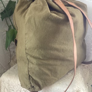 May include: A large, olive green canvas backpack with brown leather straps and a drawstring closure. The backpack is sitting on a white lace tablecloth.