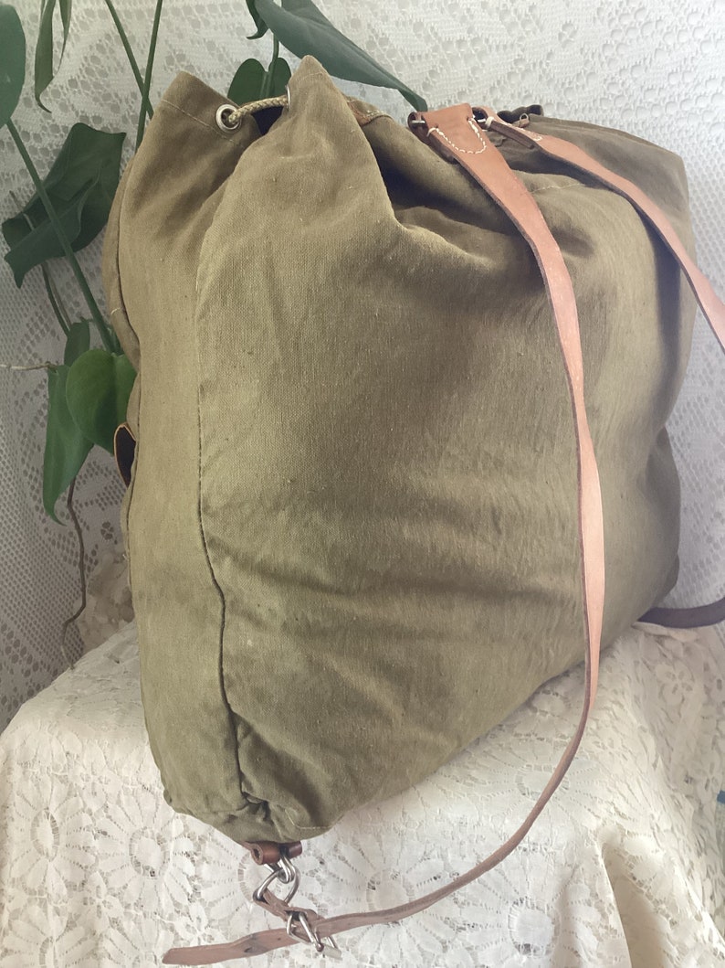 May include: A large, olive green canvas backpack with brown leather straps and a drawstring closure. The backpack is sitting on a white lace tablecloth.