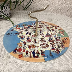 Puede incluir: Una bandeja de servir vintage con un mapa de Francia ilustrado con figuras y monumentos de dibujos animados. La bandeja tiene un asa de metal curvada y una combinación de colores azul y beige.