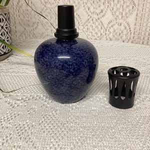 May include: A dark blue speckled ceramic essential oil diffuser with a black top and a separate black diffuser cover. The diffuser is round and sits on a white lace tablecloth. The diffuser is a home decor item.