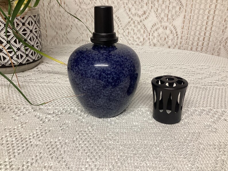 May include: A dark blue speckled ceramic essential oil diffuser with a black top and a separate black diffuser cover. The diffuser is round and sits on a white lace tablecloth. The diffuser is a home decor item.
