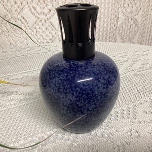 May include: A blue speckled ceramic fragrance lamp with a black top. The lamp has a rounded body and a decorative, slotted top. The background is a white, textured surface.