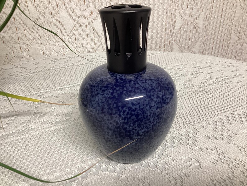May include: A blue speckled ceramic fragrance lamp with a black top. The lamp has a rounded body and a decorative, slotted top. The background is a white, textured surface.