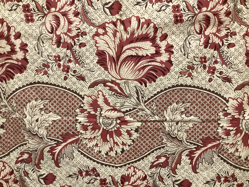 May include: A vintage fabric with a repeating pattern of red and white flowers on a cream background. The flowers have a detailed design and are surrounded by smaller floral motifs. The fabric has a woven texture and a slightly faded appearance.
