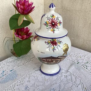 Op de afbeelding: Een witte keramische pot met deksel, versierd met bloemmotieven in rood, geel en groen. De pot heeft een geschilderde landschapsafbeelding en een goudkleurige zegel. De basis en rand zijn blauw afgewerkt.