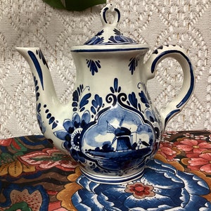 Peut inclure: Une théière en céramique blanche avec un motif bleu et blanc de Delft, représentant un moulin à vent et des motifs floraux. La théière a un couvercle et un bec.
