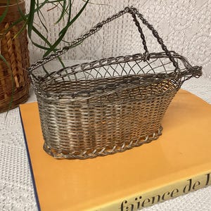 Könnte beinhalten: Ein silberfarbener Drahtkorb mit gewebtem Boden und Griff. Der Korb steht auf einem gelben Buch. Ein gewebter brauner Korb und eine grüne Pflanze befinden sich im Hintergrund.