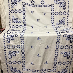 May include: White tablecloth with a blue embroidered border and scattered bird motifs. The border features floral and geometric patterns. The birds are stylized and add a whimsical touch to the classic design. This tablecloth is suitable for a dining table.