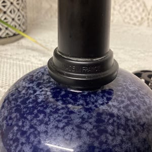May include: A blue and white speckled glass bottle with a black top. The base of the black top reads "MADE IN FRANCE". A black object is visible to the right of the bottle.