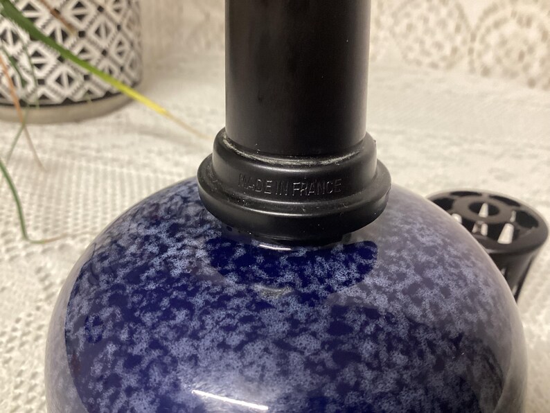 May include: A blue and white speckled glass bottle with a black top. The base of the black top reads "MADE IN FRANCE". A black object is visible to the right of the bottle.