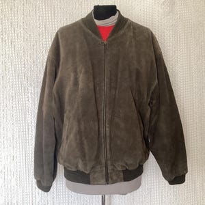 Peut inclure: Veste de bombardier en daim marron avec fermeture éclair intégrale. La veste a un col, des poignets et un ourlet côtelés. La veste est présentée sur un mannequin sur fond blanc.