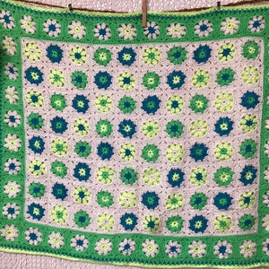 Peut inclure: Une couverture en crochet avec un fond rose et un motif de fleurs vertes, bleues et jaunes. La couverture a une bordure verte avec des fleurs blanches.