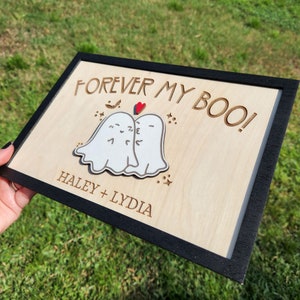May include: A wooden sign with a black frame, featuring two white ghosts with red hearts, and the text "FOREVER MY BOO! HALEY + LYDIA".