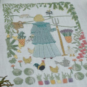 Puede incluir: Un textil de lino blanco con una escena de jardín bordada. El diseño incluye una figura con una horca, rodeada de plantas, animales y flores. El romero verde y las hojas de laurel enmarcan el textil.
