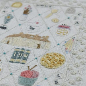 May include: Embroidered white fabric with a detailed design featuring various objects and the year "2005". The design includes a house, books, food items, and other decorative elements, all stitched in muted colors. The edges are finished with a delicate, scalloped border.