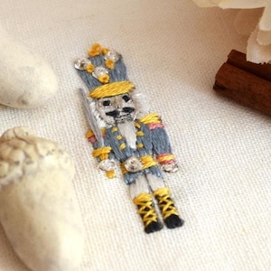 May include: A hand-stitched nutcracker embroidery with a gray uniform, yellow accents, and a white hat. The embroidery is on a white fabric background. The text "Matchbox Embroidery" is visible in the background.