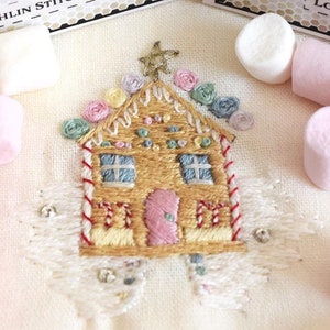 Op de afbeelding: Een witte stof met een geborduurd peperkoekhuis met snoepdetails. Het huis is omringd door roze en witte marshmallows.