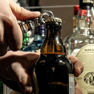 May include: A person's hand using a silver bottle opener to open a brown glass bottle of beer. The bottle has a label with text on it. The bottle is in front of a shelf with other bottles of alcohol.