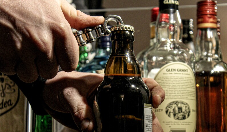 May include: A person's hand using a silver bottle opener to open a brown glass bottle of beer. The bottle has a label with text on it. The bottle is in front of a shelf with other bottles of alcohol.