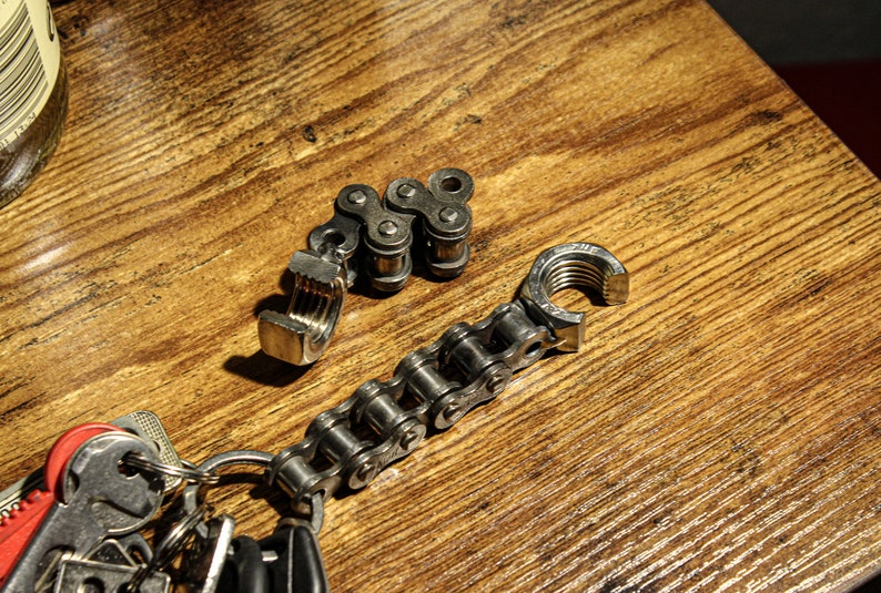 May include: A silver chain keychain with a bottle opener and a clamp attached. The keychain is on a wooden surface.