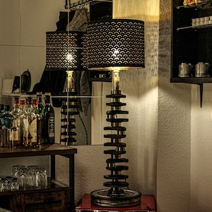 Könnte beinhalten: Eine schwarze Lampe mit einem geometrisch gemusterten Schirm steht auf einem roten Sockel. Die Lampe besteht aus einem Stapel von Metall-Motorteilen. Die Lampe ist eingeschaltet und strahlt ein warmes Licht aus.
