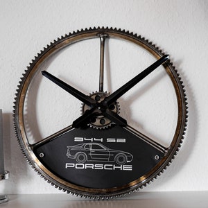 May include: A black and silver metal clock with a gear design. The clock face features a black and white silhouette of a Porsche 944 with the text "944 62 PORSCHE".