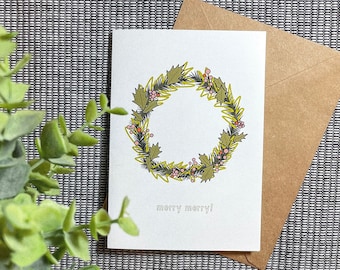 May include: A white card with a green and yellow wreath with pink berries and the text "merry merry!" printed on it. The card is sitting on a brown envelope.