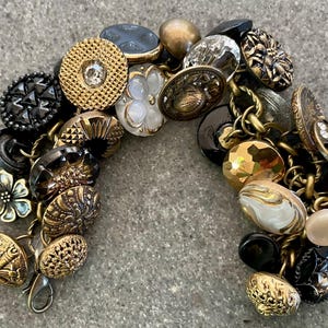May include: A vintage button bracelet featuring a variety of antique buttons in gold, black, and white. The buttons are connected by a gold-tone chain and clasp. The buttons have various textures and designs.