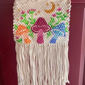 May include: A woven wall hanging featuring three colorful mushrooms in orange, magenta, and blue, with green foliage and a crescent moon. The artwork is framed by a wooden dowel and has long, cream-colored fringe.