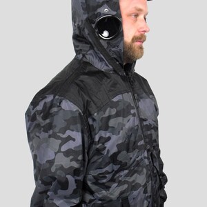May include: A black and gray camouflage jacket with a black hood and a black zipper. The jacket has a black patch on the left side of the chest with a black circle in the center.